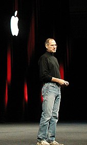 Full-length portrait of man about 50 years old wearing jeans and a black turtleneck shirt, standing in front of a dark curtain with a white Apple logo