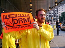Man in Tyvek suit holding a "Eliminate DRM" sign