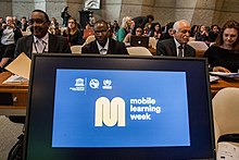 A computer screen at the front of a room of policymakers shows the Mobile Learning Week logo
