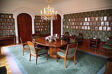 A room with pictures on the walls. In the middle of the room there is a wooden table with chairs around it.