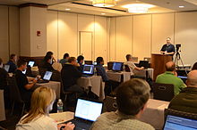 A man speaking from a podium to a room of people with laptop computers